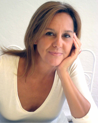 maria-duenas-escritoria Retrato de María Dueñas. Apoya su cara en su mano izquierda, con el codo sobre la mesa. Mirada amables. Viste una camiseta blanca de cuello de pico con media manga.